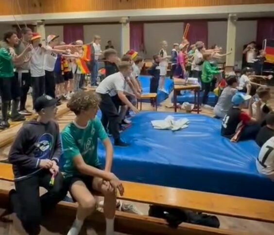 Ein Foto vom Public Viewing bei der EM 2024, auf dem viele Kinder in der HNT Vereinshalle sitzend und stehend ein Spiel verfolgen.