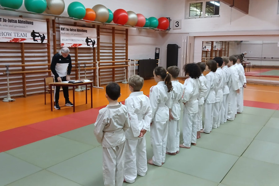 Ein Foto von den Prüflingen, die nebeneinander in einer Reihe auf der Matte stehen und Prüfer Michael Richter zuhören,