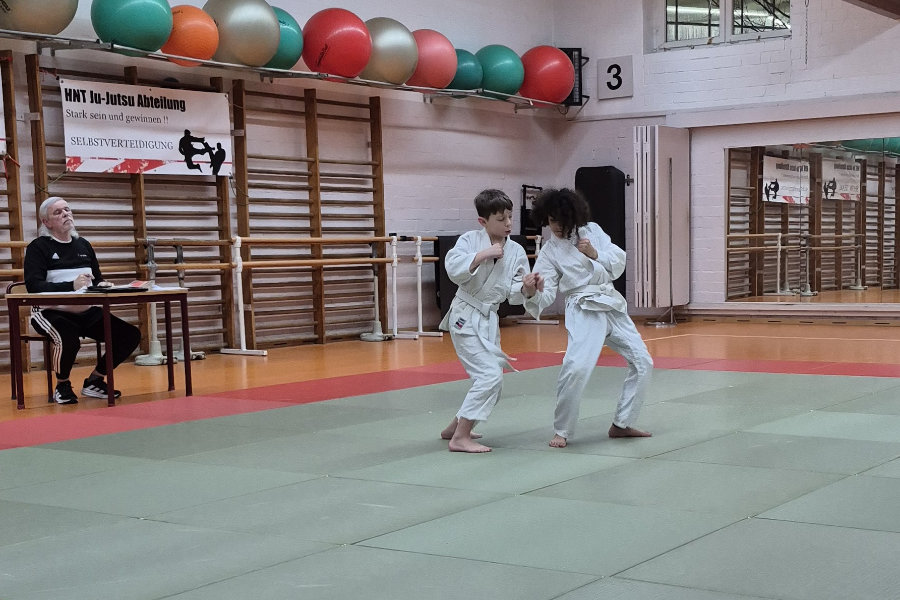 Ein Foto von zwei Prüflingen, die eine Übung auf der Matte zeigen; beide stehen dabei und halten sich gegenseitig fest.