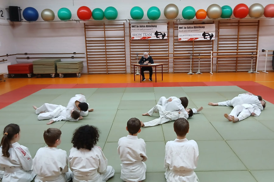 Ein Foto von der Prüfung, auf dem die Kinder paarweise Bodentechniken vorführen, ein Teil der Prüflinge schaut dabei vom Mattenrand zu.