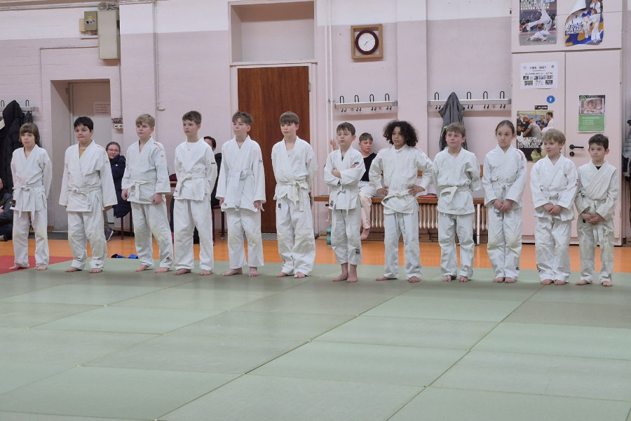 Ein Foto der Kinder, die an der Gurtprüfung teilnehmen. Sie stehen aufgereiht nebeneinander am Rand der Matte.