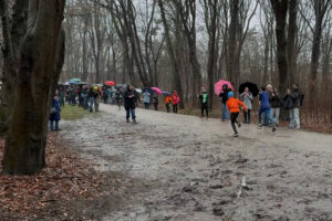 Ein Foto von der schlammigen Laufstrecke beim "Swim & Run" im Volkspark, zwei Teilnehmer laufen mit dem Rücken zur Kamera und am Rand stehen zahlreiche Zuschauer mit Regenschirmen.