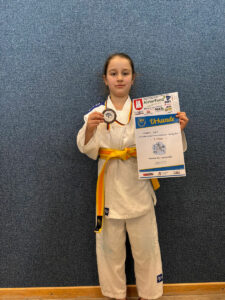 Ein Foto von Viktoria, auf dem sie alleine vor einer Wand steht und ihre Bronzemedaille und ihre Urkunde nach oben hält.