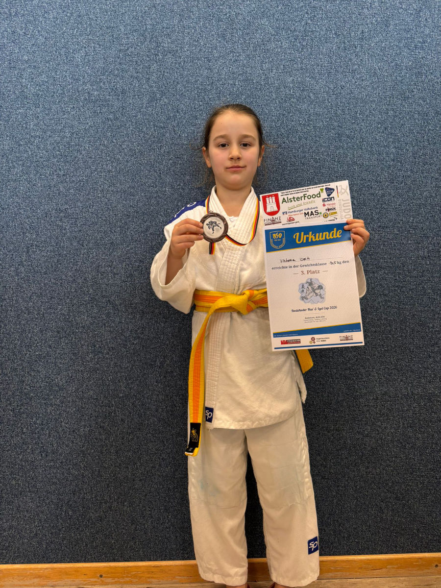 Ein Foto von Viktoria, auf dem sie alleine vor einer Wand steht und ihre Bronzemedaille und ihre Urkunde nach oben hält.