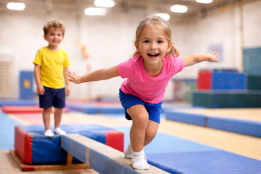 Ein Symbolfoto für das Kinderturnen, auf dem ein Mädchen auf einem Balken steht, sich nach vorne beugt und in die Kamera lächelt.
