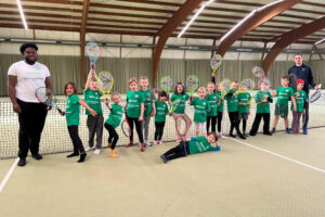 Ein Foto von Kindern des HNT-Feriencamps, die in der Tennishalle aufgereiht nebeneinander am Netz stehen und ihre Schläger freudig nach oben halten.