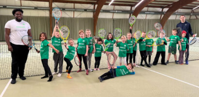 Ein Foto von Kindern des HNT-Feriencamps, die in der Tennishalle aufgereiht nebeneinander am Netz stehen und ihre Schläger freudig nach oben halten.