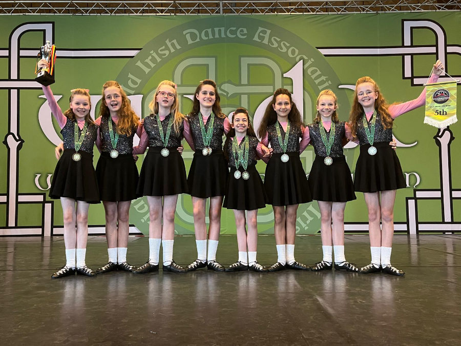 Ein Gruppenfoto des achtköpfigen Céilí-Teams der HNT, auf dem die Tänzerinnen in einer Reihe nebeneinander auf der Bühne stehen und ihre Medaillen und Trophäen präsentieren.