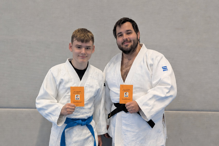 Ein Foto von Max und David, auf dem beide nebeneinander auf der Judomatte stehen und ihre Kampfrichterscheine in die Höhe halten.