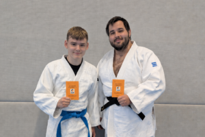 Ein Foto von Max und David, auf dem beide nebeneinander auf der Judomatte stehen und ihre Kampfrichterscheine in die Höhe halten.