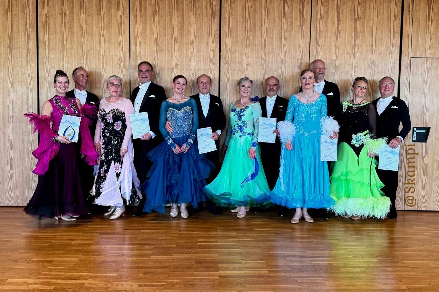 Ein Foto von Caro und Volker, die neben anderen Tanzpaaren stehen und ihre Trophäe und Urkunde präsentieren.