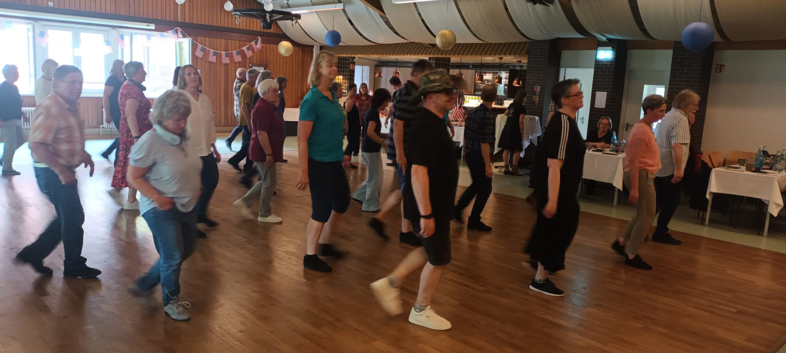 Ein Foto vom Tanztee der Gruppe "Just For Fun", auf dem im Saal viele Teilnehmende in Reihen vor- und hintereinander tanzen.