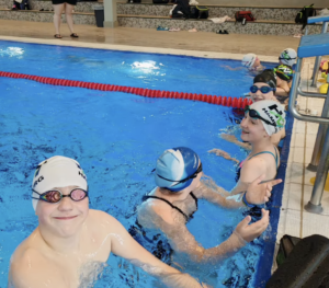 Ein Foto von mehreren HNT-Schwimmer:innen, die im Schwimmbecken am Rand unterhalb der Startblöcke warten.