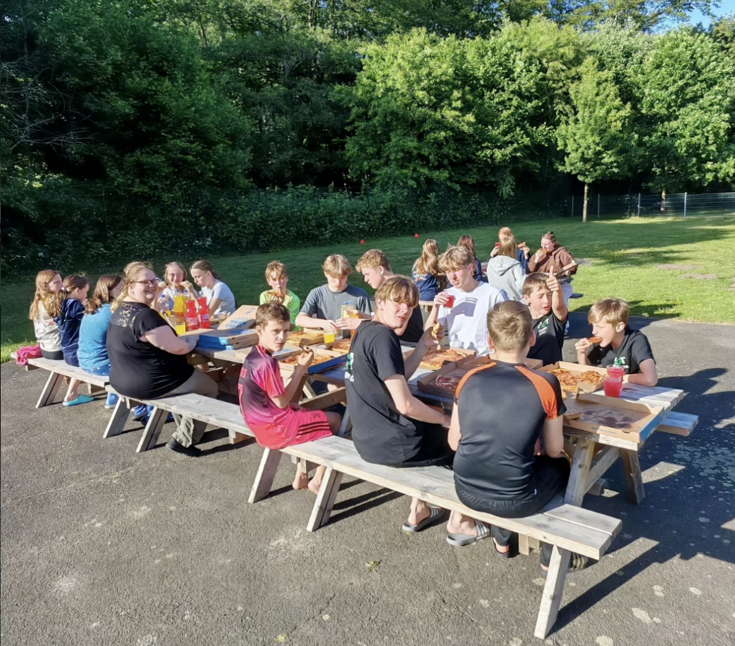 Kurztrainingslager HNT Schwimmen Mai 2026 Ein Foto von der HNT-Trainingsgruppe, die an mehreren Holztischen im freien sitzt und eine Essenspause macht.