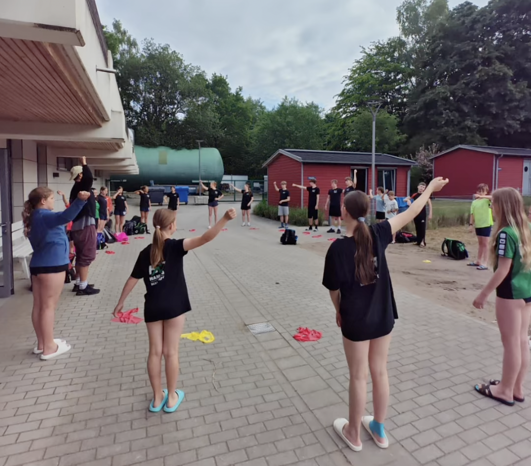 Kurztrainingslager HNT Schwimmen Mai 2026 Ein Foto von der HNT-Trainingsgruppe, die in einem großen Kreis vor einem Gebäude steht und eine Übung macht.