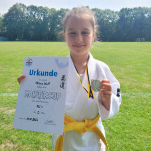 Ein Foto von Viktoria, die draußen auf einer Wiese steht und ihre Urkunde und die Medaille für den zweiten Platz hochhält.