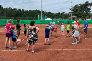 Ein Foto von Teilnehmenden, die auf einem Tennisplatz im Kreis stehen und einem Trainer zuhören.