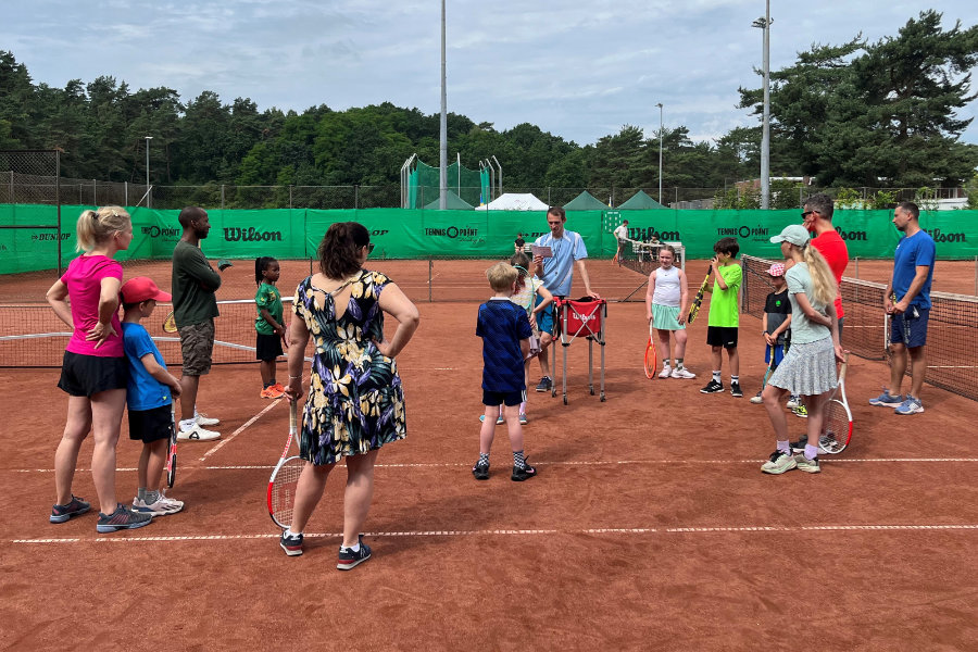 Ein Foto von Teilnehmenden, die auf einem Tennisplatz im Kreis stehen und einem Trainer zuhören.