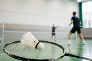 Ein Symbolfoto zum Thema Badminton, das einen Badmintonschläger und einen -ball im Vordergrund zeigt, dahinter unscharf zwei Badmintonspieler in Aktion.