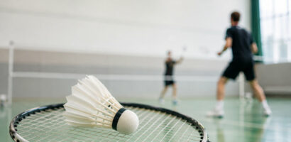 Ein Symbolfoto zum Thema Badminton, das einen Badmintonschläger und einen -ball im Vordergrund zeigt, dahinter unscharf zwei Badmintonspieler in Aktion.