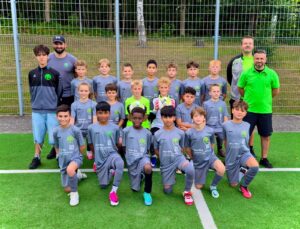 Die HNT 1.D steht gemeinsam mit ihrem Trainerteam für ein Mannschaftsfoto auf dem Fußballplatz.