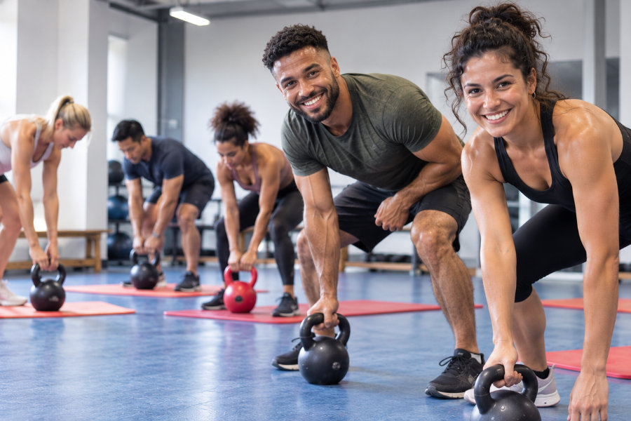 Ein mit KI erstelltes Foto, das mehrere Personen beim Training mit Kugelhanteln in einer Sporthalle zeigt, zwei Personen lächeln dabei freundlich in die Kamera.